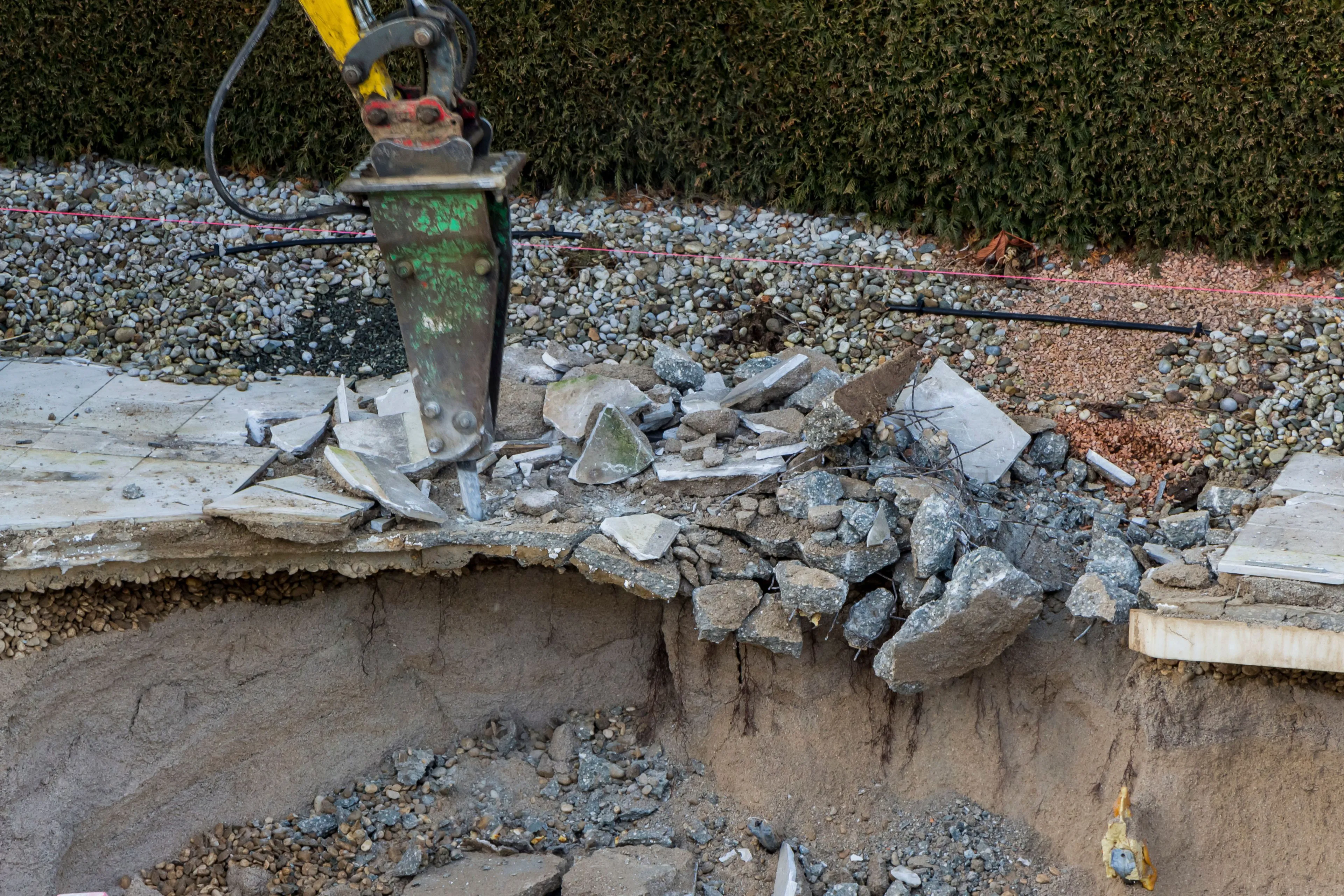A hydraulic jackhammer attachment on an excavator breaking through the thick concrete shell of an inground swimming pool during a full removal project in Dallas
